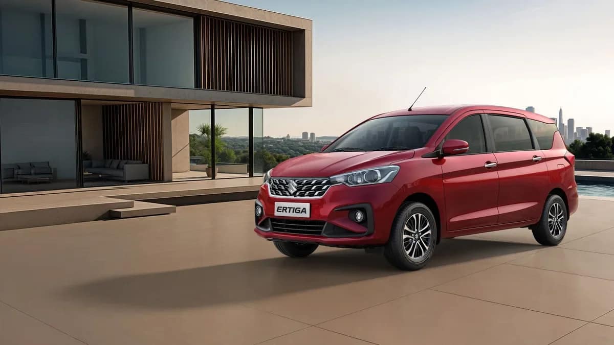 Red Maruti Suzuki Ertiga MPV parked outside a modern glass house, front three-quarter view